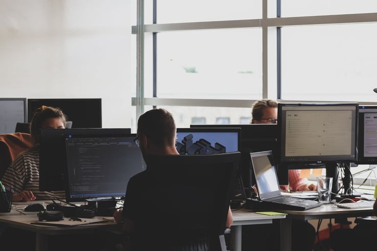 A person coding in front of a monitor. Surrounded by desks and computers. 